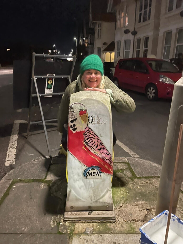 Mew Welch crouching behind a bollard with her Budgie paste up