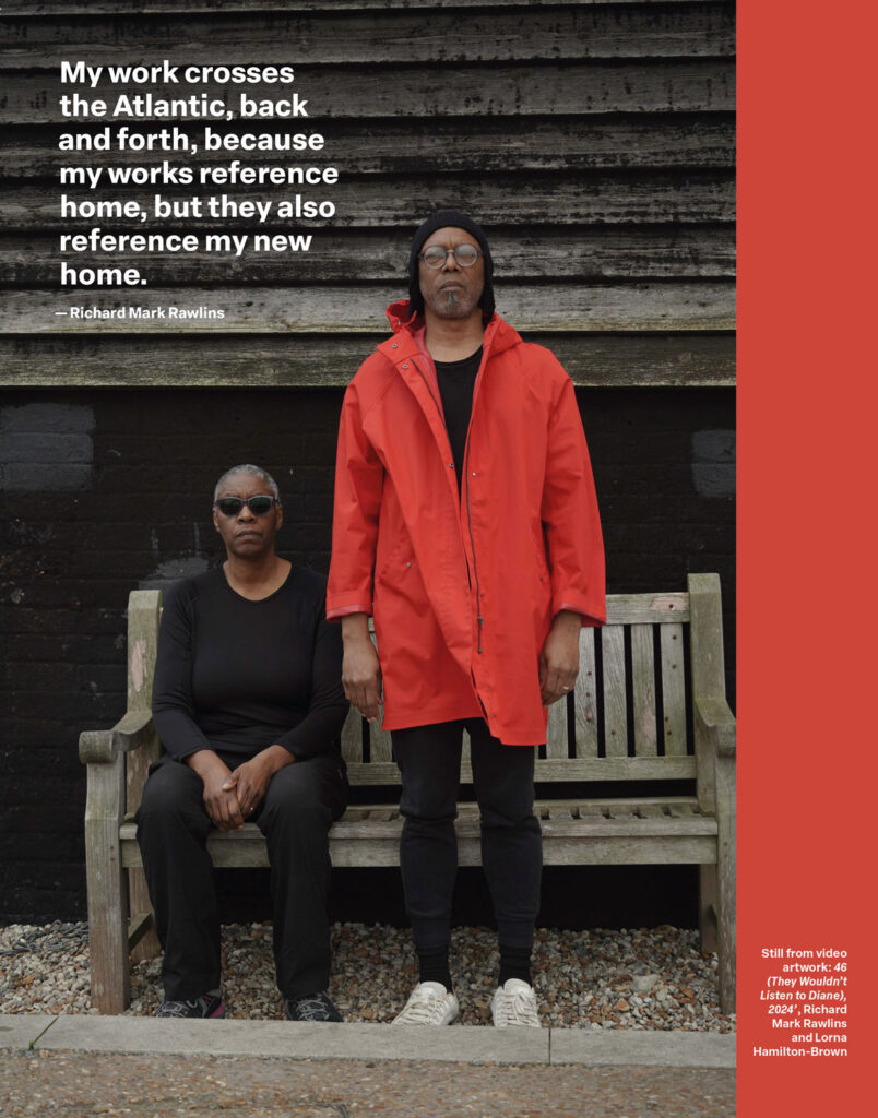 Richard Mark Rawlins in a red coat is standing in front of a bench with fellow artist Linda Hamilton Brown MBE seated.